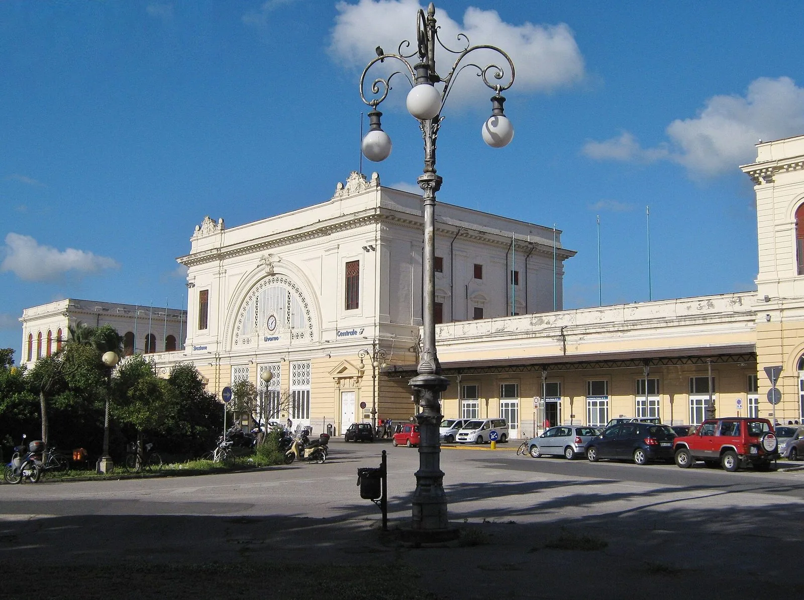 La facciata chiara della stazione di Livorno Centrale con il grande finestrone ad arco, un lampione in ghisa in primo piano e il piazzale sotto un cielo azzurro