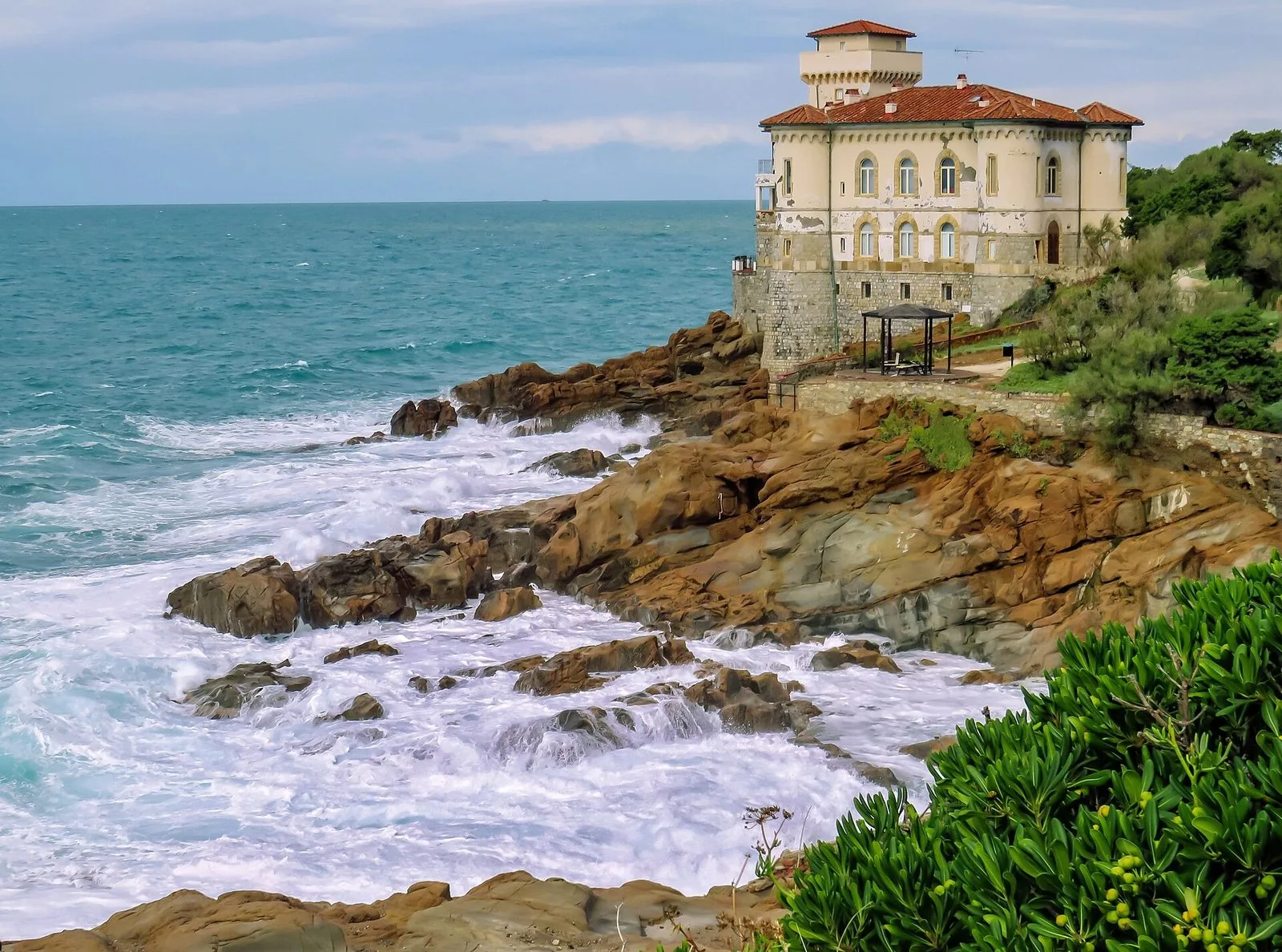Mare mosso con onde bianche che si infrangono sugli scogli di arenaria sotto il Castello del Boccale, una villa color crema con torre e tetti rossi affacciata sul mare, con piante verdi in primo piano