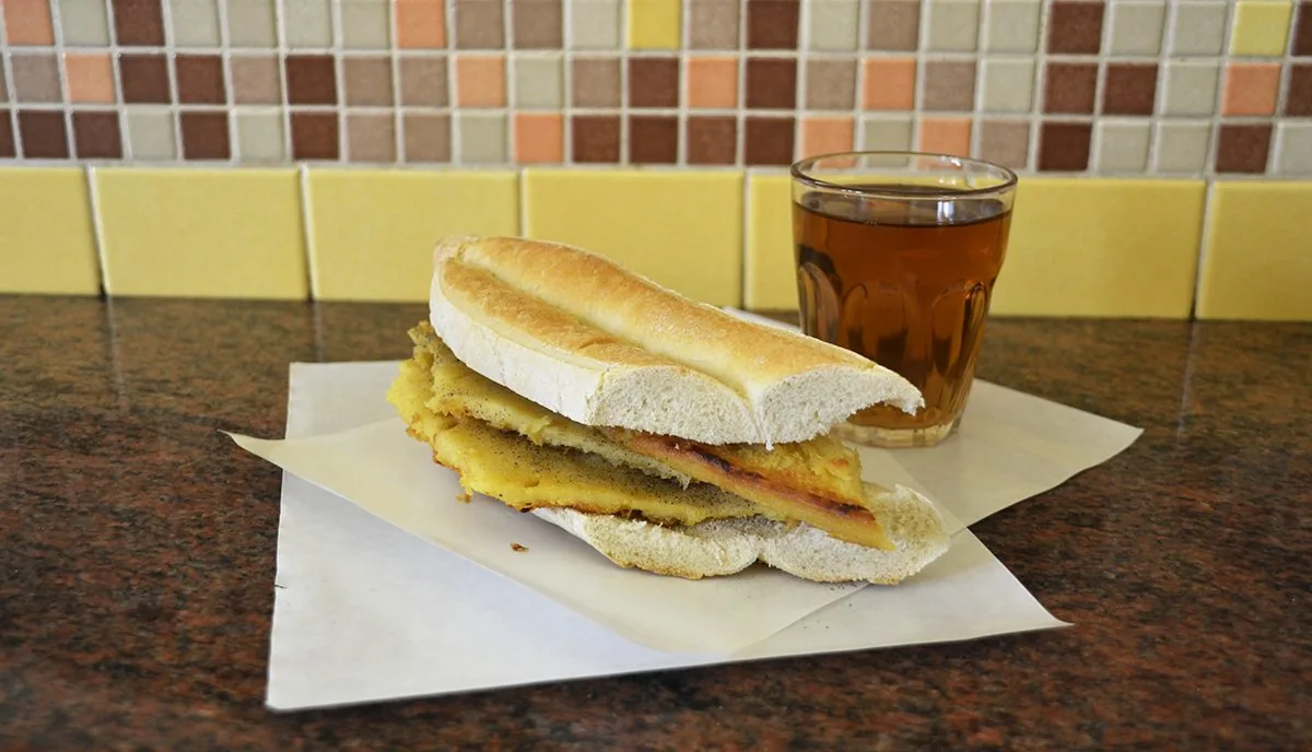 Un 5 e 5: panino di pane bianco farcito con strati di torta di ceci dorata e pepata, su carta da forno, accanto a un bicchiere con una bibita ambrata